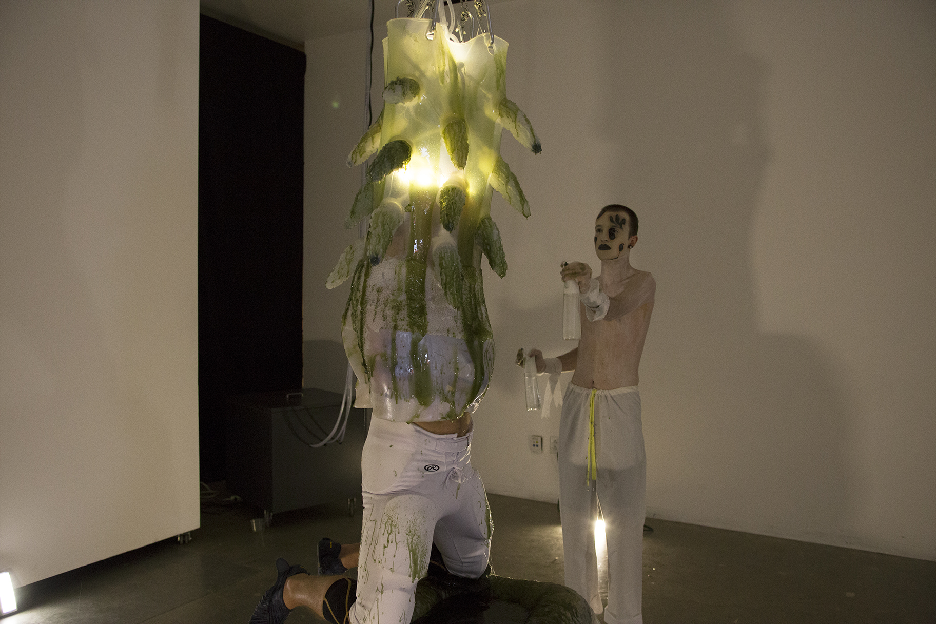 A man engulfed in a gelatenous form covered in tentacles knealing over a pool of green slime while a shirtless man painted in white sprays him with moisture